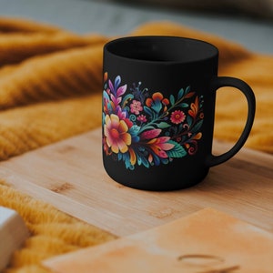 May include: Black ceramic mug with a colorful floral design. The flowers are pink, yellow, orange, and blue. The mug is on a wooden surface.