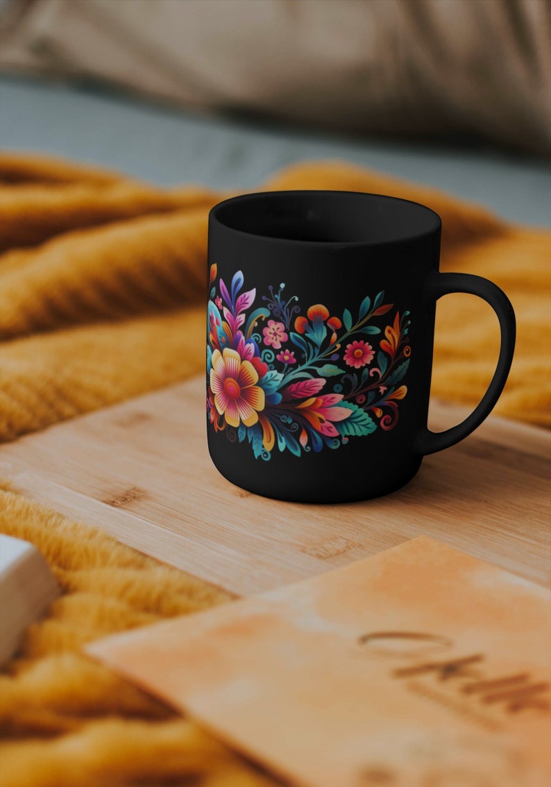 May include: Black ceramic mug with a colorful floral design. The flowers are pink, yellow, orange, and blue. The mug is on a wooden surface.