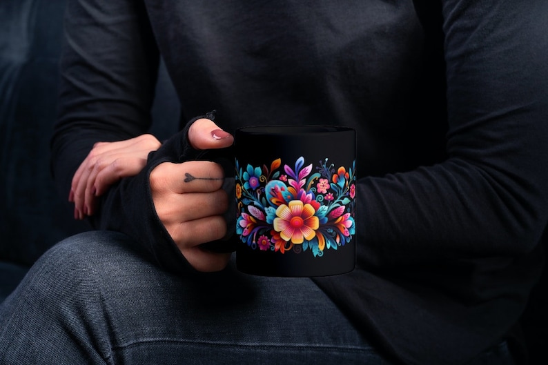 May include: Black coffee mug with a colorful floral design. The flowers are pink, yellow, blue, and green. The mug is being held by a person wearing a black shirt and jeans.