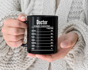 Op de afbeelding: Een zwarte koffiemok met het woord "Doctor" in witte letters. De mok heeft een voedingswaardelabel met waarden voor hard werken, toewijding, passie, respect, kennis, cafeïne en spijt. De mok wordt vastgehouden.