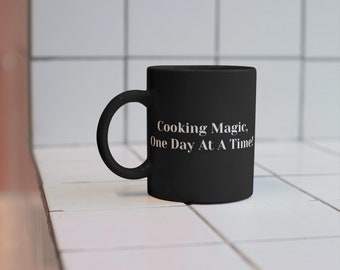 May include: Black ceramic mug with white text that reads "Cooking Magic, One Day At A Time!" The mug is sitting on a white tiled surface.