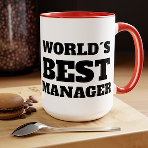 May include: White ceramic mug with a red rim and handle. The mug has black text that reads "WORLD'S BEST MANAGER".