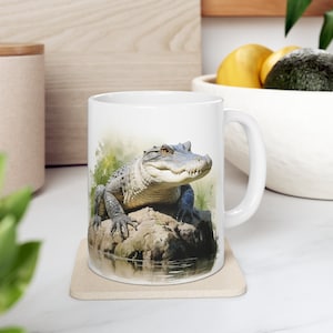 May include: White ceramic mug featuring a colourful illustration of an alligator on a rock in a swamp. The alligator is facing the viewer with its mouth closed.