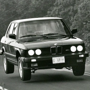 May include: A black BMW saloon car, photographed in monochrome, appearing to be in motion. The car is shown mid-air, with its wheels off the road. Headlights are on, and a number plate is visible.