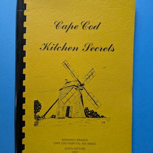 Könnte beinhalten: Ein Vintage-Kochbuch mit dem Titel "Cape Cod Kitchen Secrets" mit gelbem Einband. Das Cover zeigt eine Schwarz-Weiß-Illustration einer Windmühle und Text. Das Buch ist mit einer schwarzen Spiralbindung gebunden.