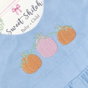 May include: A light blue baby dress with embroidered pumpkins. The dress has ruffles around the neckline and sleeves. The text "Sweet Shiloh Baby + Child" is on a pink tag.