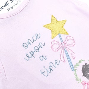 May include: Pink baby onesie with the text "Once upon a time" embroidered in blue and pink thread. A blue and pink wand with a yellow star on top is embroidered on the onesie. The text "Sweet Shiloh Baby + Child" is on a white tag.