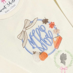 Puede incluir: Body de bebé color crema con un diseño bordado. El diseño presenta un círculo azul con un lazo, iniciales, calabazas, hojas de otoño y una porción de tarta. La etiqueta dice "Sweet Shiloh Baby + Child."