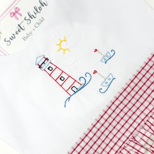 May include: White baby dress with a red and white gingham skirt. The bodice of the dress features an embroidered lighthouse, sun, and sailboats.