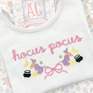 Pode incluir: Um macacão branco para bebê com o texto bordado em rosa "hocus pocus" e duas bruxas voando em vassouras com estrelas e luas.
