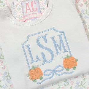 Peut inclure: Une grenouillère blanche avec un monogramme bleu "LSM" dans un cadre décoratif et deux citrouilles orange brodées avec des tiges et des feuilles vertes.