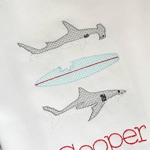 May include: White embroidered shirt with two sharks and a surfboard, the name "Cooper" is embroidered in red below the sharks.