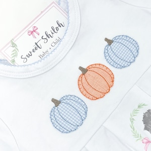 Pode incluir: Macacão branco para bebé com três abóboras bordadas. As abóboras são de xadrez azul e branco, laranja e de xadrez azul e branco. O macacão tem uma bainha com babados e um decote festoneado.