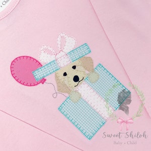 May include: A pink long-sleeved shirt with a light blue and white gingham gift box with a brown dog peeking out of the top. The dog is holding a pink balloon. The shirt has the text "Sweet Shiloh Baby + Child" on the top left corner.
