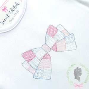 Puede incluir: Un body blanco para bebé con un lazo de cuadros rosa y azul bordado en la parte delantera. El body tiene una pequeña etiqueta en la parte delantera que dice "Sweet Shiloh Baby + Child".