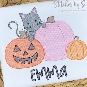 May include: A white onesie with an embroidered design of a gray cat sitting on top of a jack-o'-lantern pumpkin. There are two other pumpkins, one pink and one orange, in the background. The name "EMMA" is embroidered below the pumpkins.