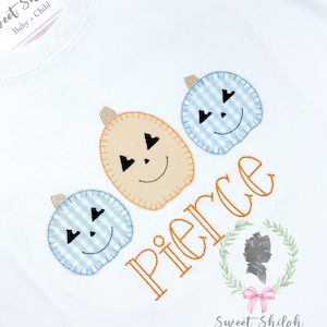May include: White onesie with three embroidered pumpkins and the name "Pierce" in orange thread. The pumpkins are blue and white gingham, orange, and yellow. The onesie has a label that says "Sweet Shiloh Baby + Child".