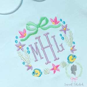 May include: A light blue shirt featuring an embroidered wreath of sea life, including purple starfish, yellow fish, green seaweed, and shells, surrounding the purple monogram letters MHL, topped with a green bow and the text Sweet Shiloh Baby + Child.