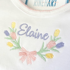 Pode incluir: Macacão branco para bebê com uma coroa de flores bordada em rosa e o nome "Elaine" em bordado roxo.