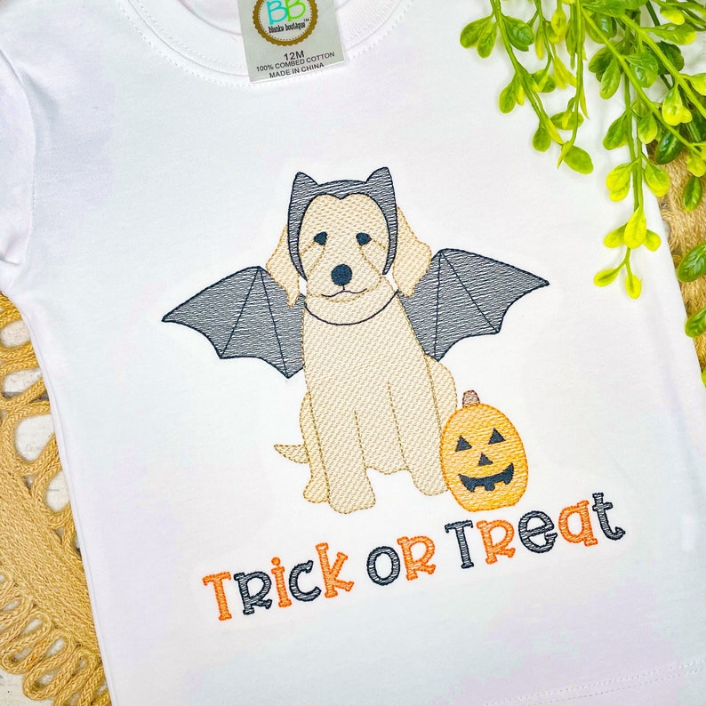 K&ouml;nnte beinhalten: Ein wei&szlig;es T-Shirt mit einer Stickerei eines hellen Hundes, der schwarze Fledermausfl&uuml;gel und einen Haarreif mit Fledermausohren tr&auml;gt. Neben dem Hund befindet sich ein orangefarbener K&uuml;rbis. Der Text "TRICK OR TREAT" ist in Orange und Schwarz.