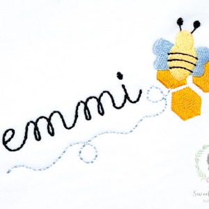 Peut inclure: Broderie avec le nom "emmi" en écriture noire, accompagnée d'une abeille jaune et noire avec des ailes bleues et des formes de nid d'abeilles. Le tout sur fond blanc.