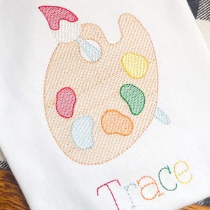 May include: White fabric embroidered with a painter's palette and brush, featuring colourful paint splotches. The name "Trace" is embroidered in rainbow colours below the palette. The design is stitched with a textured effect.