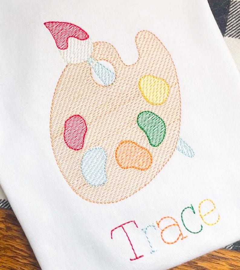 May include: White fabric embroidered with a painter's palette and brush, featuring colorful paint splotches. The name "Trace" is embroidered in rainbow colors below the palette. The design is stitched with a textured effect.