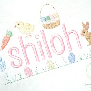 Peut inclure: Broderie avec le nom "Shiloh" en rose, ornée de motifs de Pâques. Comprend une carotte, un poussin, un panier d'œufs, un lapin et des œufs décorés. Sur fond blanc avec de l'herbe verte.