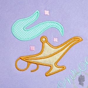May include: Embroidered design featuring a gold genie lamp with a cream-colored body and a teal, gingham-patterned smoke effect. Pink star accents are scattered around the lamp on a lavender background.