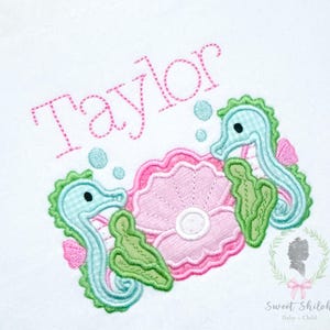 May include: Embroidered design with two seahorses, a seashell, and the name "Taylor" in pink thread. The seahorses are light blue with pink accents, and the seashell is pink. Green seaweed and blue bubbles complete the design.