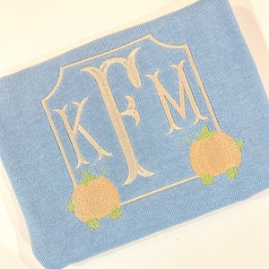 May include: A light blue knitted blanket with a white embroidered monogram "KFM" in a decorative frame. Two embroidered orange pumpkins with green stems are on either side of the monogram.
