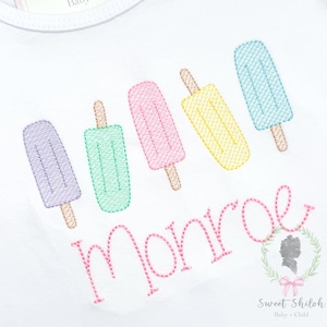 Pode incluir: Body de bebé branco bordado com cinco desenhos coloridos de picolés em roxo, verde, rosa, amarelo e azul. O nome "Monroe" é bordado em rosa abaixo. Um logotipo com o texto "Sweet Shiloh Baby + Child" também é visível.