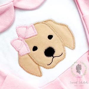 May include: A light pink baby outfit with a white panel featuring an embroidered golden retriever puppy face with a pink bow. The puppy has black eyes and nose, and the outfit has a collar and sleeves.