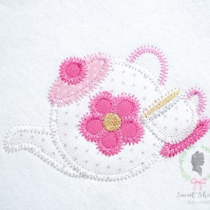 Könnte beinhalten: Gestickte weiße Teekanne mit rosa Blume und Akzenten. Die Teekanne hat einen rosa Griff und Ausguss sowie eine rosa Blume mit goldenem Zentrum. Das Design ist auf einem weißen Hintergrund mit kleinen grauen Punkten gestickt. Der Text "Sweet Shiloh Baby + Child" befindet sich unten rechts.