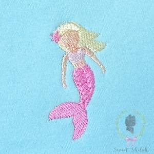 May include: An embroidered patch featuring a mermaid with blonde hair, a pink flower accessory, a light purple top, and a pink tail, stitched onto a light blue fabric background. A small logo with the text Sweet Shiloh is in the bottom corner.