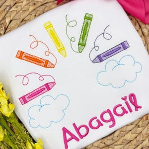 Könnte beinhalten: Weißer Stoff mit gestickten bunten Buntstiften und Wolken. Der Name "Abagail" ist rosa gestickt. Die Buntstifte sind orange, gelb, grün, rosa und lila, mit geschwungenen Linien.