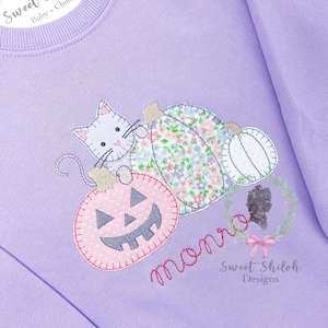 May include: A purple sweatshirt with an embroidered design of a cat, pumpkins, and the word "monroe". The pumpkins are pink, white, and blue with floral patterns. The design is surrounded by a wreath of leaves and a pink bow.