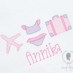 May include: Pink and purple embroidered travel-themed design featuring a suitcase, airplane, and swimwear. The name "Annika" is embroidered in pink script. The design is on a white background.
