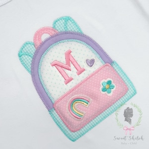 May include: A colorful, embroidered backpack design in pastel colors. The backpack features a large letter "M", a rainbow, a flower, and a heart. The design is on a white background and is likely for a child.