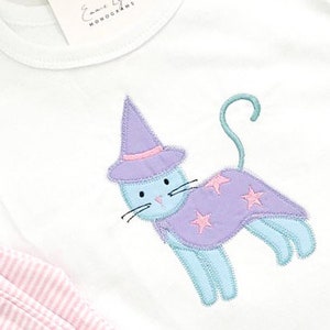 Puede incluir: Una camiseta blanca con un gato azul que lleva un sombrero de bruja morado y una capa morada con estrellas rosas. El gato está bordado en la camiseta.