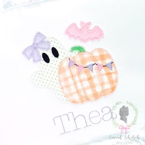 May include: A white fabric item featuring an embroidered design of a ghost with a lavender bow, a plaid orange pumpkin with a green stem and small bunting, and a pink bat. The name Thea is embroidered in purple below the ghost and pumpkin.