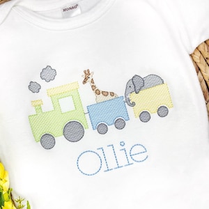 Puede incluir: Un pelele blanco con un diseño bordado de un tren verde con un vagón amarillo, una jirafa en el vagón del medio y un elefante en el último vagón. El tren tiene ruedas grises y el pelele está personalizado con el nombre "Ollie" en hilo azul.