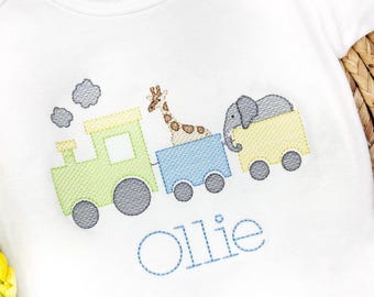Peut inclure: Une grenouillère blanche avec un motif brodé d'un train vert avec un wagon jaune, une girafe dans le wagon du milieu et un éléphant dans le dernier wagon. Le train a des roues grises et la grenouillère est personnalisée avec le nom "Ollie" en fil bleu.