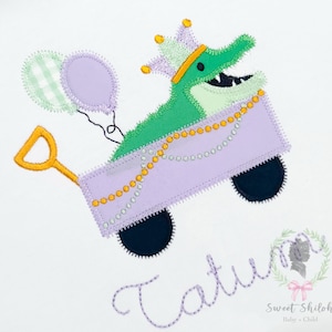 May include: An embroidered design featuring a green alligator wearing a crown, riding in a purple wagon. The wagon is decorated with gold beads and pulled by a yellow handle. Two balloons float above the wagon. The name "Tatum" is stitched below.