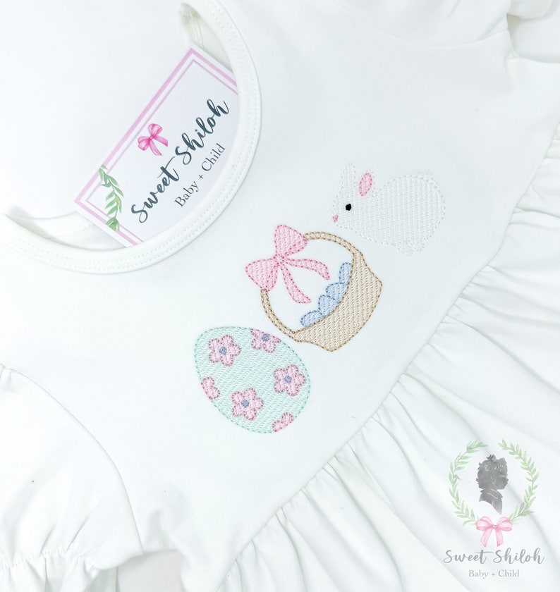 K&ouml;nnte beinhalten: Wei&szlig;es Babykleid mit gestickten Ostereiern, einem Korb mit Schleife und einem Hasen. Das Kleid hat einen R&uuml;schen-Saum und einen runden Ausschnitt. Der Text "Sweet Shiloh Baby + Child" befindet sich auf einem Etikett, das am Kleid befestigt ist.