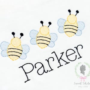 Puede incluir: Tela blanca bordada con tres abejas amarillas y negras con alas azul claro. El nombre "Parker" está bordado en escritura negra debajo de las abejas. Un pequeño logotipo con texto se encuentra en la esquina inferior derecha.