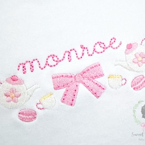 Puede incluir: Diseño bordado con un lazo rosa, tazas de té, teteras y macarons. El nombre "Monroe" está escrito en rosa en cursiva. El diseño está sobre un fondo blanco. El diseño es de Sweet Shiloh Baby & Child.