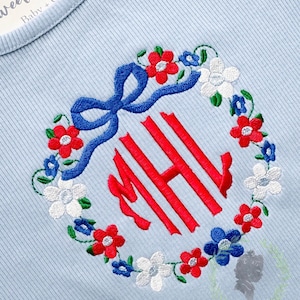 Puede incluir: Tejido acanalado azul claro con una corona floral bordada. La corona presenta flores rojas, blancas y azules, que rodean un monograma rojo. Un lazo azul está en la parte superior. El texto "Sweet St" es visible en la parte superior.