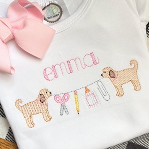 Può includere: Maglietta bianca a maniche corte con il nome "emma" ricamato in rosa. Due cani marroni ricamati tengono una ghirlanda con materiale scolastico. Un grande fiocco rosa è attaccato alla maglietta.