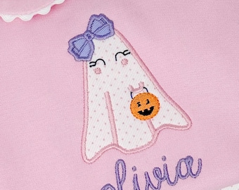 May include: Pink Halloween-themed garment featuring an embroidered ghost with a purple bow, holding a jack-o'-lantern. The ghost has a polka-dot pattern. The name "olivia" is embroidered in purple script below the ghost. The garment has a white scalloped collar.